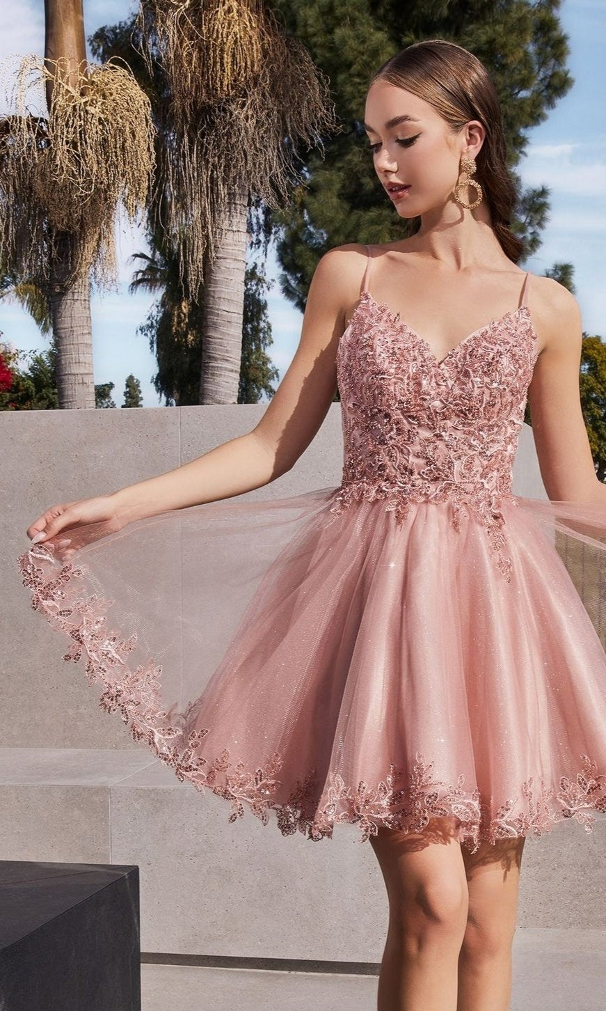 A young woman stands outdoors in the Short Homecoming Dress CD0213 with embroidery, holding the side of her pink, lace-embellished sleeveless skirt, with trees and concrete structures in the background.